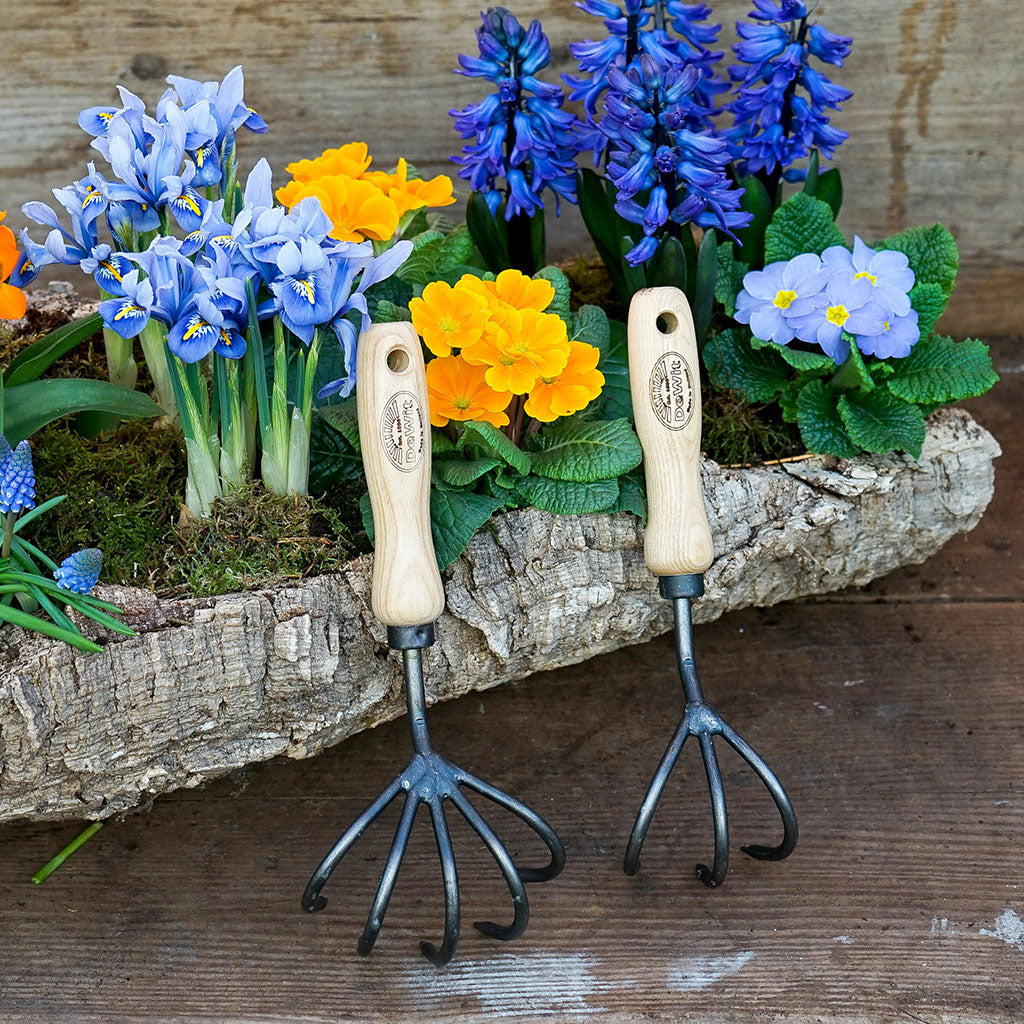 Two DeWit handheld cultivators displayed in front of a variety of flowers in a stone planter.