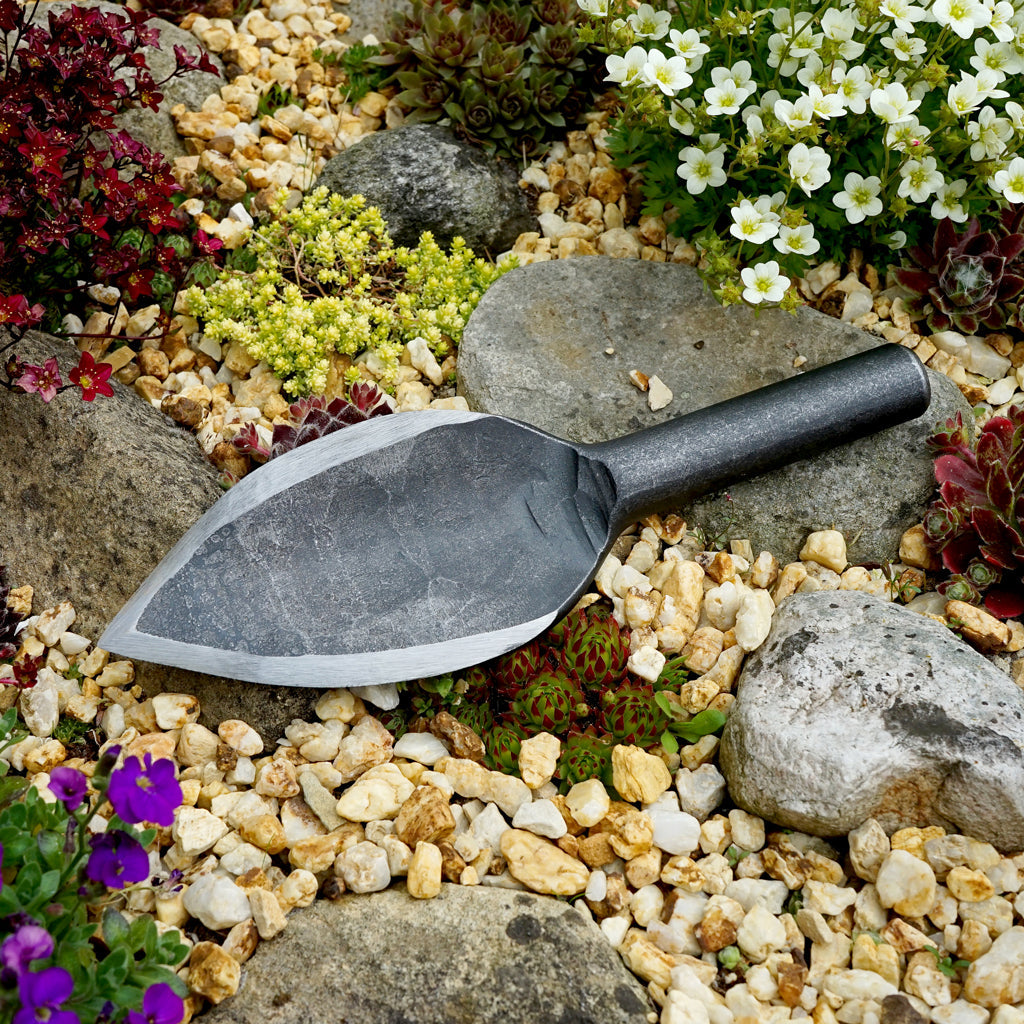 DeWit's heavy-duty, Bad Ass Garden Trowel, made from one piece of hand-forged, recycled boron steel displayed in a rock garden. 