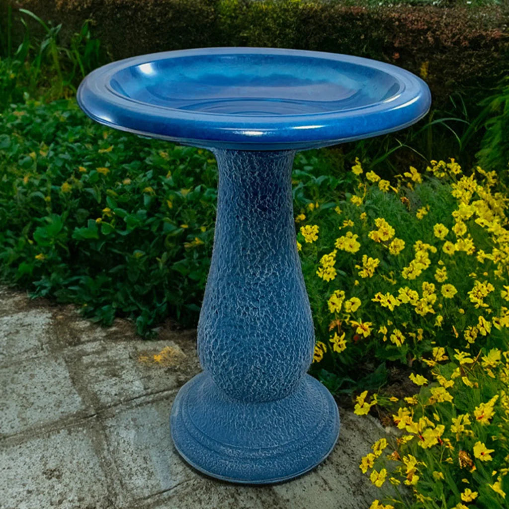 Brilliant blue pedestal birdbath on a patio next to flowering plants.